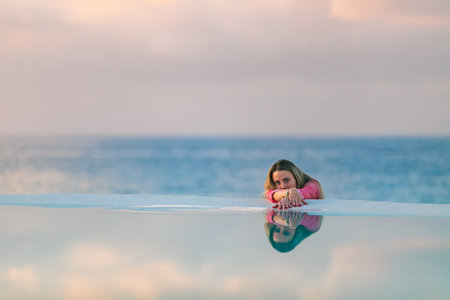A young girl in a red shirt, at dawn by the sea pool, calm and relaxed looking at the reflection in...