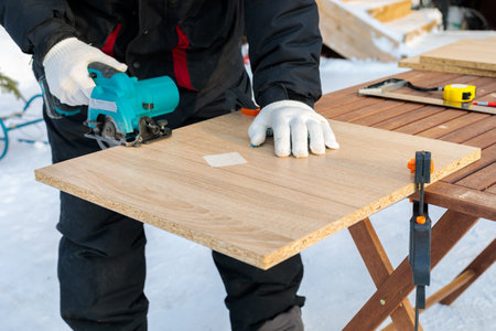 A male worker cuts a wooden panel attached to a workbench with clamps using a circular disc on a battery.の写真素材