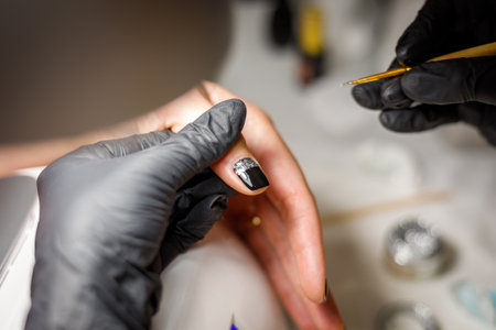 In a beauty salon, the master does a manicure, applies a pattern to the nail with a brush, black varnish with a golden pattern.の写真素材