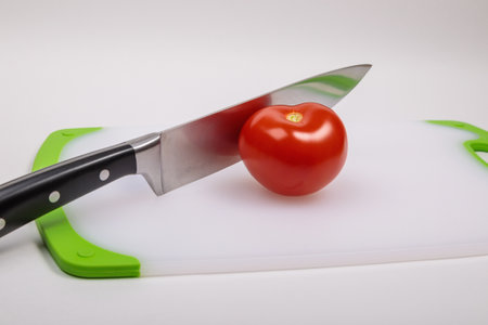 A fresh red tomato and a large chef's knife lie on a cutting board. On an isolated white background.の写真素材