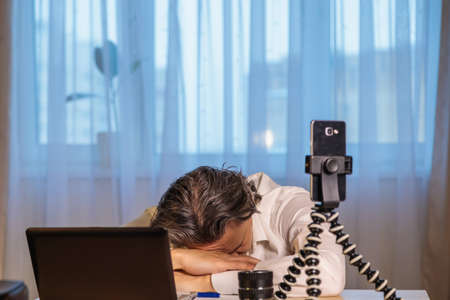 A 50-year-old blogger is recording an online seminar on his smartphone for his subscribers. With the help of modern devices, a computer and a smartphone, the master class is broadcast live. The coach shows different emotions.の写真素材