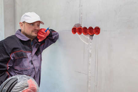 An electrician stands against the wall with an electrical cable for sockets.の写真素材
