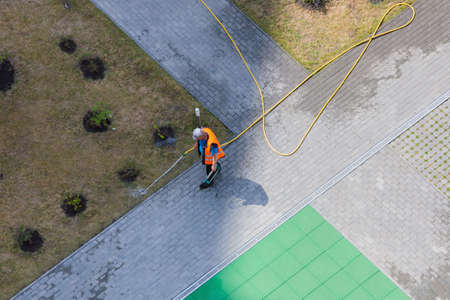 A worker is watering the bushes in the yard with a hose from the hot sun.