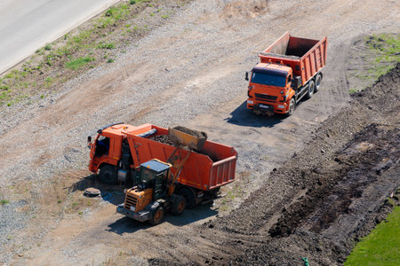 A worker on a tractor with a bucket loads earth into a truck. Chelyabinsk, Russia, May 15, 2021のeditorial素材