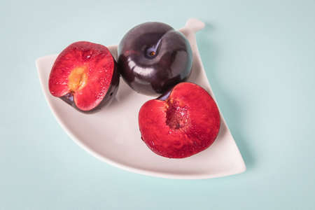 On a white saucer is a large plum, cut in half, and next to it is a whole plum.
