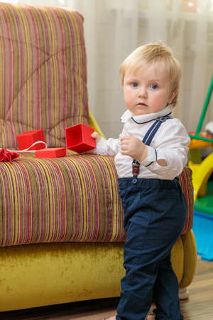 Little boy, child stands by the couch with a festive gift box. In a nice outfit, a white shirt and trousers with suspenders.の写真素材