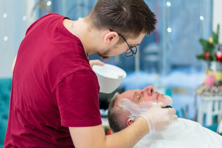 A master applies shaving cream to the beard of an adult man in a hairdressing salon with a shaving brush. Shave your beard in the salon. Moscow, Russia November 17, 2016のeditorial素材
