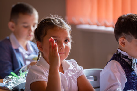 The first grader sits at her desk with her hand raised, wants to answer the teacher.