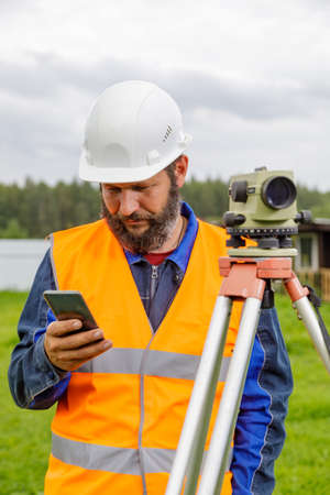 A civil engineer with an optical level looks into a mobile phone.の写真素材