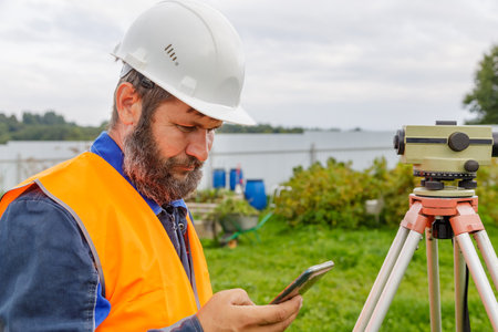 A civil engineer with an optical level looks into a mobile phone. A bearded man is looking for information on his mobile phone.の写真素材