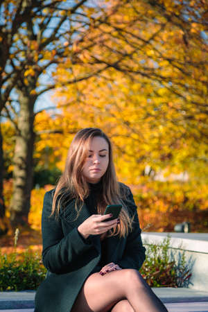 A woman with long hair sits on a bench in an autumn park and looks into a mobile phone. Autumn...