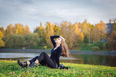 A beautiful girl poses while sitting on the grass by a pond in an autumn parkの写真素材