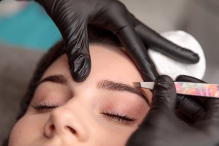 Beautician using tweezers to shape the eyebrows of a young woman in a beauty salon