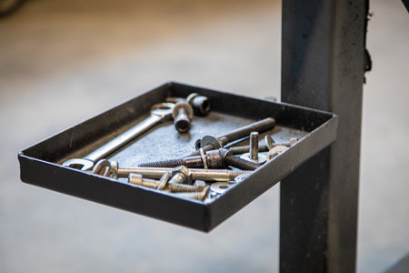 Metal tray holding various screws, bolts, nuts, and a wrench, attached to a stand in a garage or workshop settingの写真素材