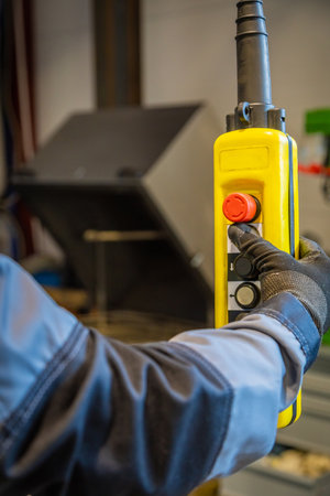 Factory worker wearing gloves using a control pendant, pushing red emergency button on yellow control panelの写真素材
