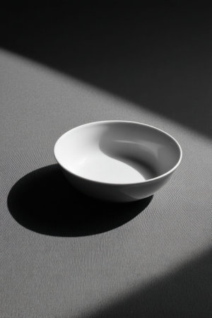 Empty white bowl casting a shadow on a gray surface under dramatic lighting, creating a minimalist and elegant compositionの素材