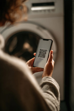 Female laundry worker scans QR code on mobile device, positioned near washing machine, highlighting the integration of technology in everyday laundry tasksの素材
