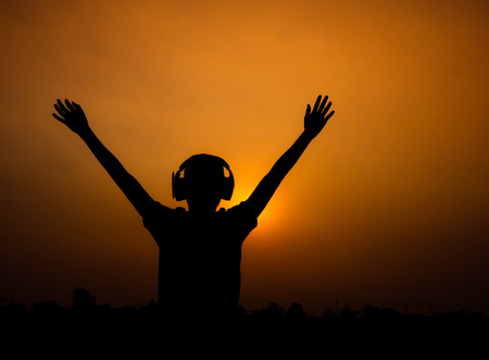 Silhouette of Asian teen boy is listening to music from headphones and raising arms at sunset evening sky background.の写真素材