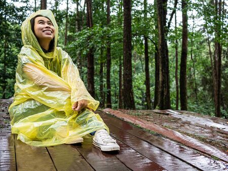 Happy Asian woman wearing yellow raincoat sit on wooden floor under rain.の写真素材