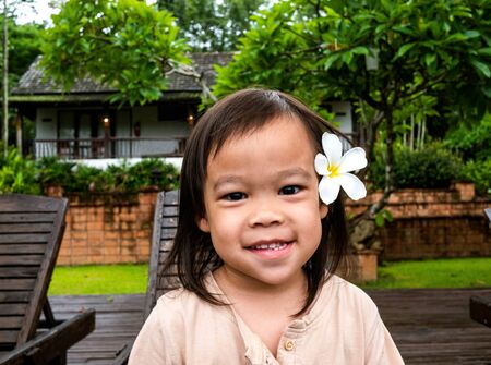 Portrait of Asian little child girl with white plumeria flower on the ear and smile face sitting on wooden chair  nearing swimming pool.の写真素材