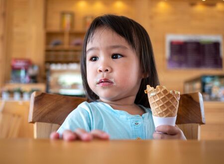 Portrait of Asian little child girl eating ice cream in cone deliciously in cafe.の写真素材