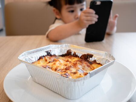 Little Asian child girl eating lunch while watching cartoon on the smartphone in a restaurant with her family. Child addicted to mobile phones.の写真素材