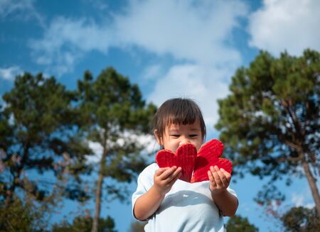 Cute little girl holding Two red heart-shaped recycled cardboard on blue sky and green tree background. Love symbol for valentines day. Concepts of Love and Romance.の写真素材
