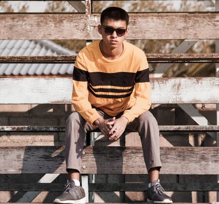 Portrait of a young handsome teenager boy in yellow long sleeve t-shirt and wear black glasses, sit on wooden stage outdoors. lifestyle people concept.の写真素材