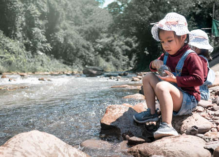 Two girls sitting on a rock by the river were having fun feeding the fish in summer day. Family travelling trip in vacation.の写真素材