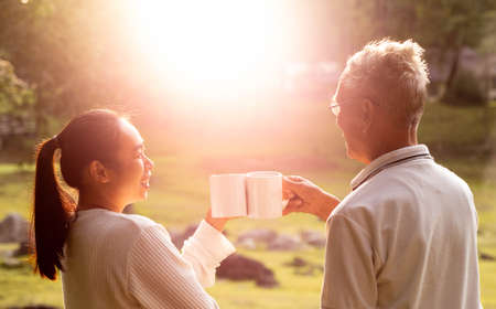 Rear view of granddaughter and grandfather drinking hot coffee outdoor and enjoying landscape during holiday on the mountain with sunlight in morning.の写真素材
