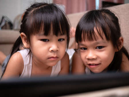 Two sibling girls lying on the sofa and watching digital tablets together in the living room.