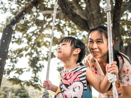 Happy mother pushing laughing daughter on swing in summer park. Family spend time together on vacation.の写真素材