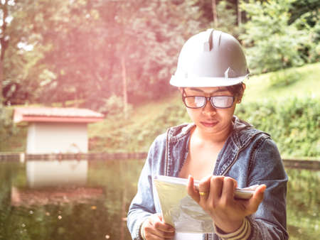 Female ecologist in safety hat working on site at the dam and stands reading the blueprints at the construction site.の写真素材