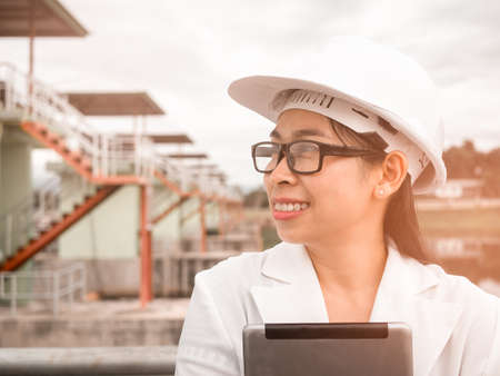 Female engineer in a white helmet talking radio communication (walkie talkie) and holding a digital tablet, stands in construction site of the hydroelectric dam.の写真素材