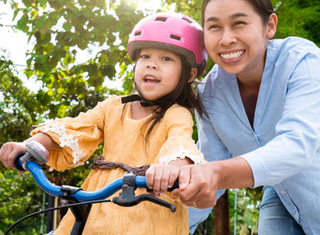 Cute little girl in safety helmet learn to ride a bike with her mother in summer park. Outdoor sports for kids. childhood happiness. family spending time together.の写真素材