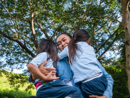 Two sibling little girls with mom laughing and hugging each other on warm and sunny summer day in...