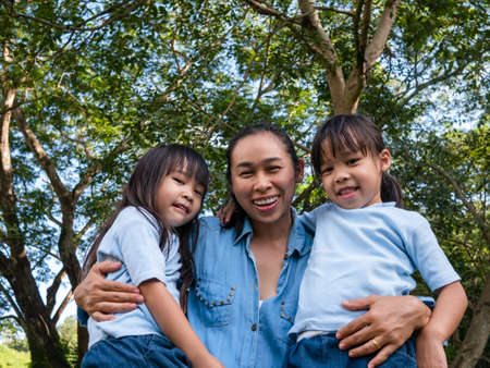 Two sibling little girls with mom laughing and hugging each other on warm and sunny summer day in...
