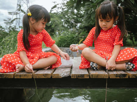 Two little sibling girls in red dress role playing fishing sitting on a wooden bridge by the pond. Childhood happiness.の写真素材