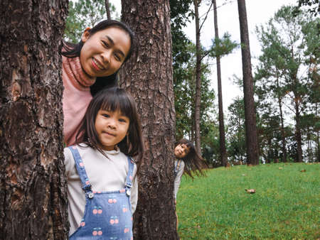 Happy mother and daughters standing behind a big tree and smiling at the camera. Happy family playing outdoors. Family spent time together.の写真素材