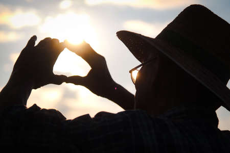 Silhouette of Senior farmer in a hat looking into the distance at sunset and making a heart shape on a golden sky background.の写真素材