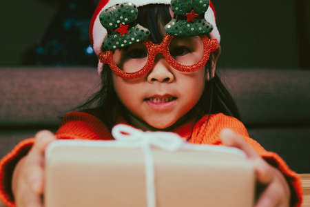 Happy little girl in Santa hat giving a Christmas present at home. Happy New Year and Merry Christmas.の写真素材