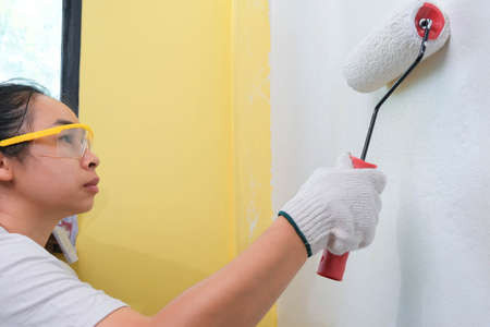 Young woman decorator wearing protective goggles paints the walls with a roller. Repair and interior...
