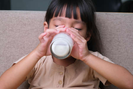 Portrait of a cute Asian little girl holding a glass of milk sitting on the sofa at home. Small girl...