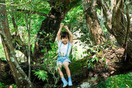 Cute little girl in boots playing in the garden, climbing a tree. Happy girl relaxing on tree branch.の写真素材