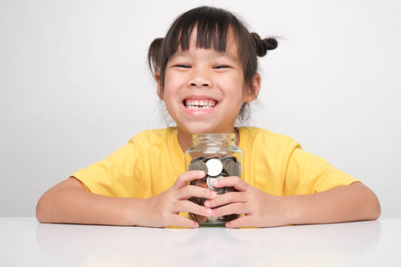 Cute Asian little girl smiling happily holding money coins in clear jar sitting at a table on white background and looking at the camera. Children learning about saving for future conceptの写真素材