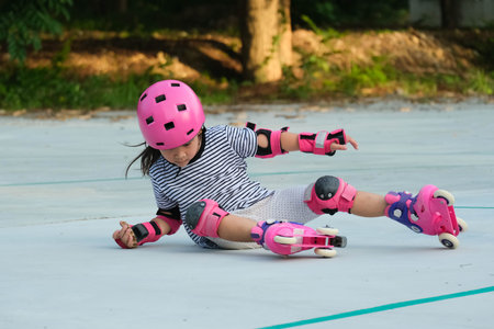 Cute Asian little girl in protective pads and safety helmet practicing roller skating in the park. Exciting outdoor activities for kids. A preschooler wearing roller skates falls down.の写真素材