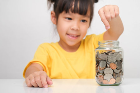 Happy smiling cute Asian girl holding a coin and putting it into a clear jar sitting at a table on a white background. Children learning about saving for future conceptの写真素材