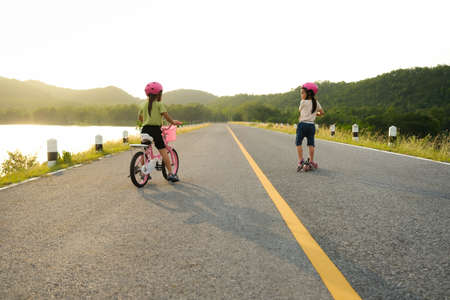 Cute little girl riding a bicycle and her sister riding a scooter on a lake road at sunset. Happy sisters doing outdoor activities together. Healthy Summer Activities for Kids.の写真素材