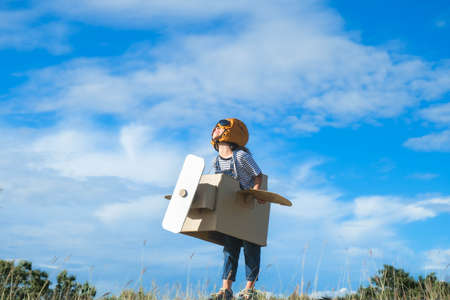 Cute dreamer little girl playing with cardboard planes in the meadow on a sunny day. Happy kid playing with cardboard plane against blue summer sky background. Childhood dream imagination concept.の写真素材