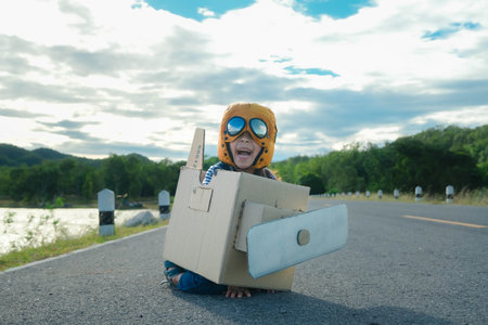 Cute dreamer little girl playing with cardboard planes on a lake road on a sunny day. Happy kid playing with cardboard plane against blue summer sky background. Childhood dream imagination concept.の写真素材